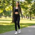 Modelo veste jaqueta cropped W One Brand Store verde militar dry fit com top e legging pretos em um parque ensolarado